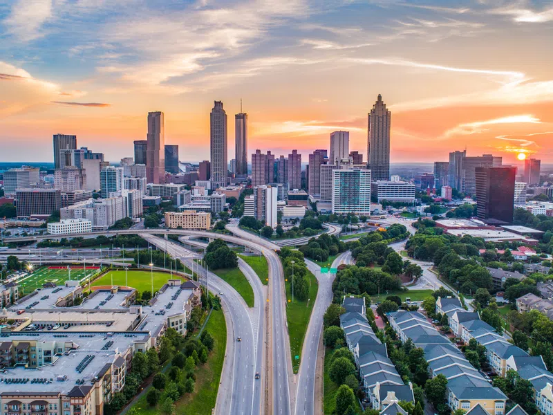 Aerial view of a city skyline at sunset with highways, green parks, and residential buildings in the foreground.