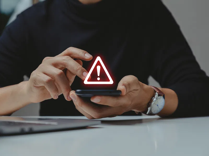 Person holding smartphone showing red warning alert icon with exclamation mark on screen.