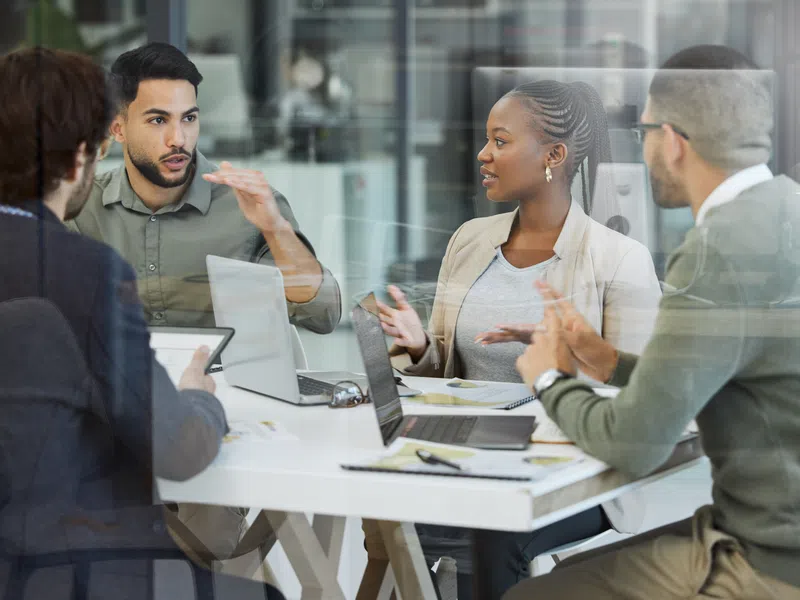 Diverse coworkers engaged in a discussion around a table with laptops and documents in a modern office.