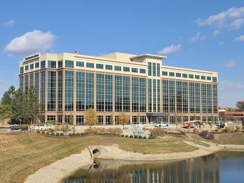 Modern glass office building reflecting blue sky beside a small pond with autumn trees and landscaping