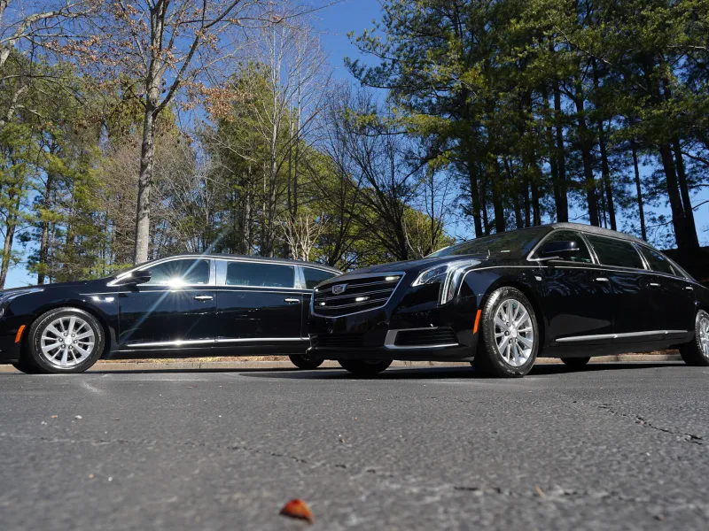 Two black Cadillac limousines parked on asphalt surrounded by trees under clear blue sky.