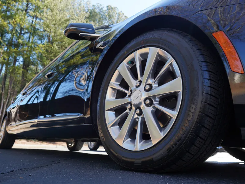 Close-up of a black car's front wheel and tire on a sunny day with trees in the background