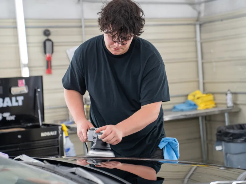 Young man in black shirt polishing a black car hood with an electric buffer in a garage workshop.