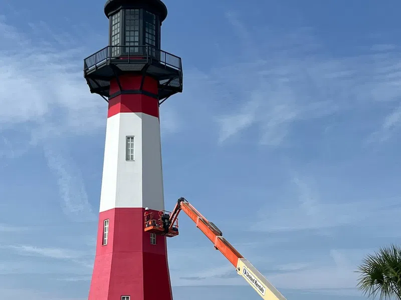 Red and white lighthouse with maintenance workers on orange lift under clear blue sky near street with traffic cones.