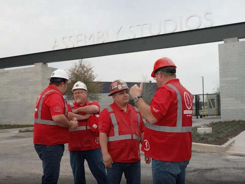 Four construction workers in red safety gear and helmets discussing outside Assembly Studios entrance on a cloudy day.