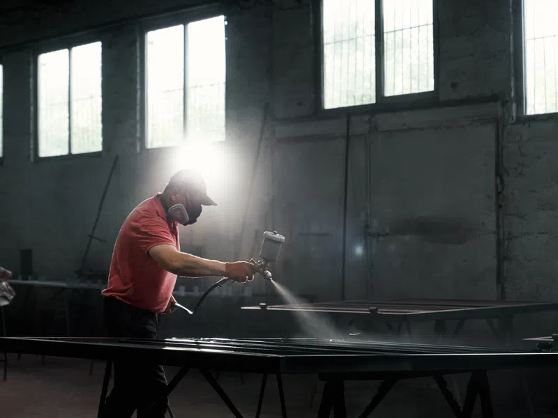 Worker wearing mask spray painting metal frames in a workshop with sunlight through windows