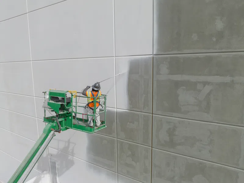 Worker in safety gear power washing a large concrete wall using a green lift platform on a construction site.