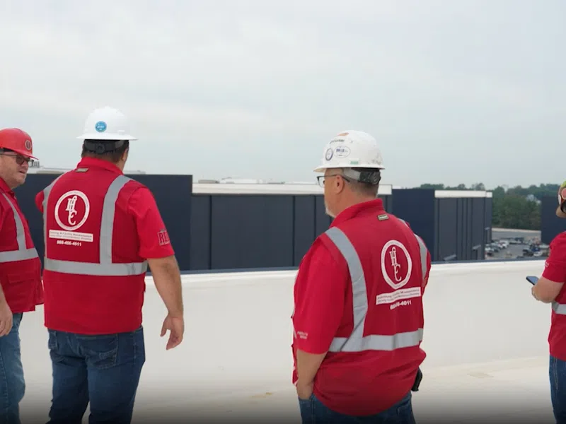 Four construction workers in red safety vests and helmets standing on a rooftop overlooking buildings.