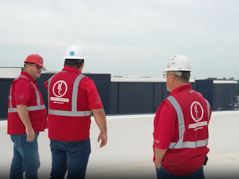 Four construction workers in red safety vests and helmets standing on a rooftop overlooking buildings.