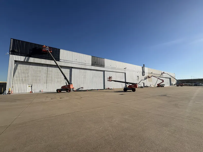Workers using boom lifts to paint a large industrial warehouse exterior under a clear blue sky.