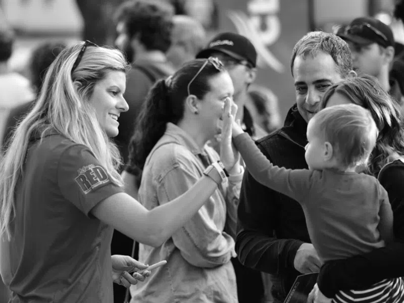 Woman in red shirt giving a high five to a child in pink shirt held by adults at an outdoor event.