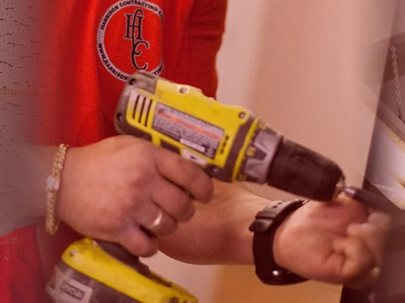 Two construction workers in red shirts using power tools to install drywall indoors wearing safety gear.