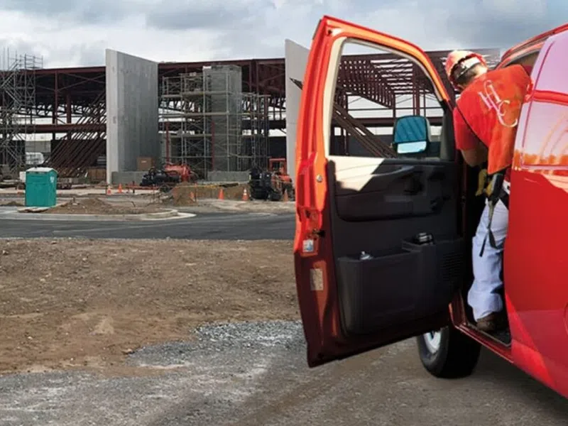 Red maintenance van parked near a construction site with building framework and worker in safety gear.