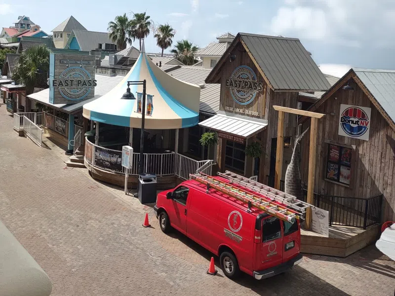 Red service van parked outside rustic seafood restaurant and shops on a sunny day with palm trees.