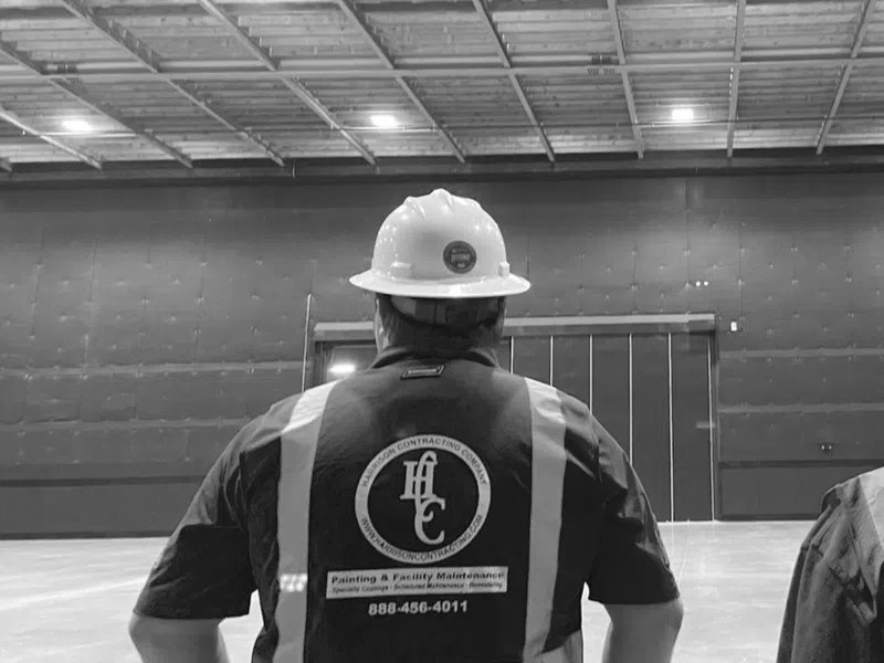Worker in red safety vest and white hard hat stands in large empty industrial warehouse with exposed wooden ceiling.