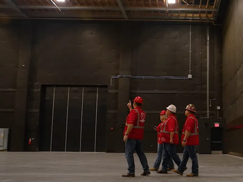 Four construction workers in red shirts and helmets inspect a large empty warehouse with concrete floors and high ceilings.