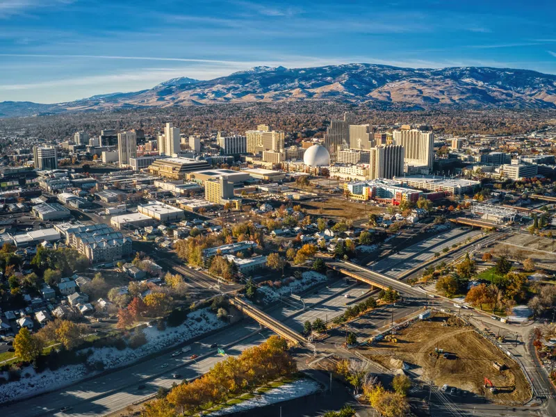 Aerial view of a cityscape with a mix of buildings, highways, and mountain range under a clear blue sky.