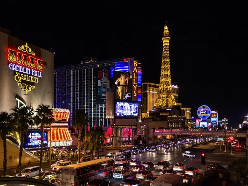 Night view of Las Vegas Strip with Bally's, Paris Eiffel Tower replica, casinos, and busy traffic with bright neon lights.