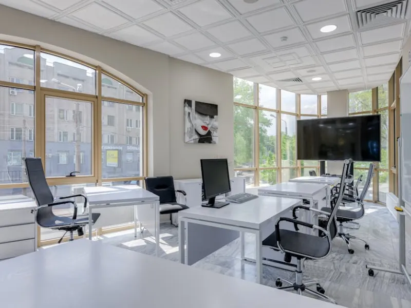 Modern bright office with white desks, black chairs, large windows, computer monitors, and a wall-mounted TV screen.