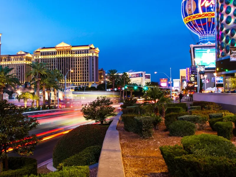 Nighttime view of Las Vegas Strip with illuminated Caesars Palace, palm trees, and traffic light trails along the road.