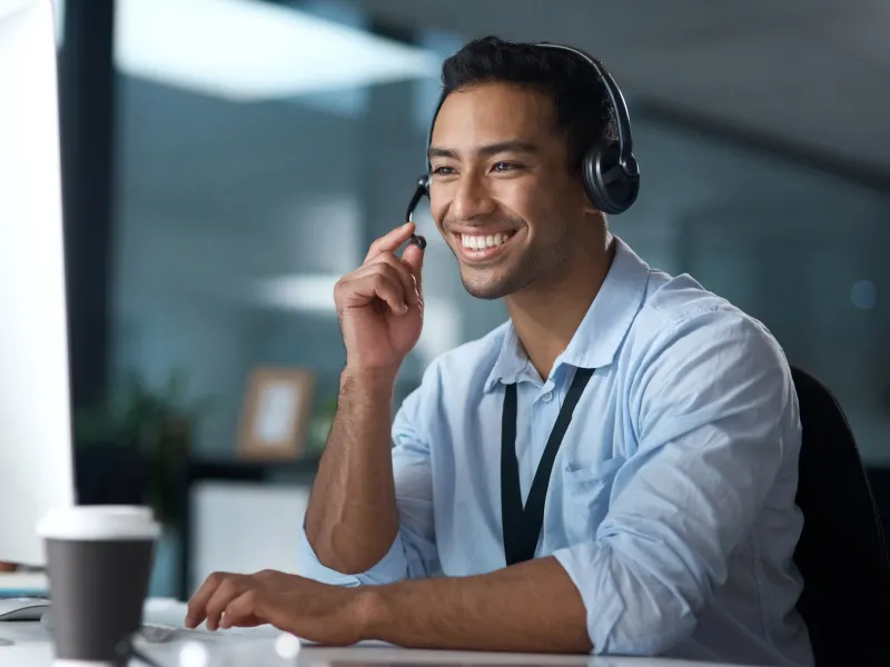 Smiling man wearing headphones at a computer, engaged in a call, working in a modern office.