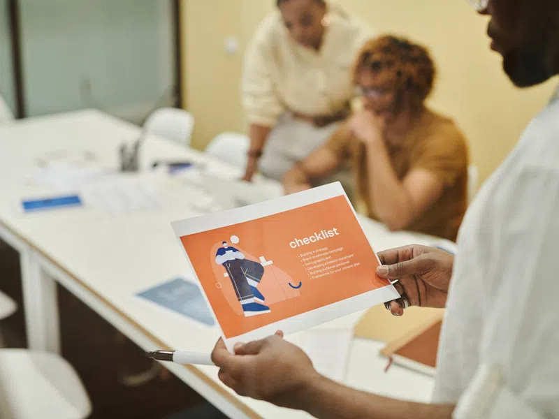 Person holding an orange checklist paper in a meeting with two colleagues at a white table with notebooks.