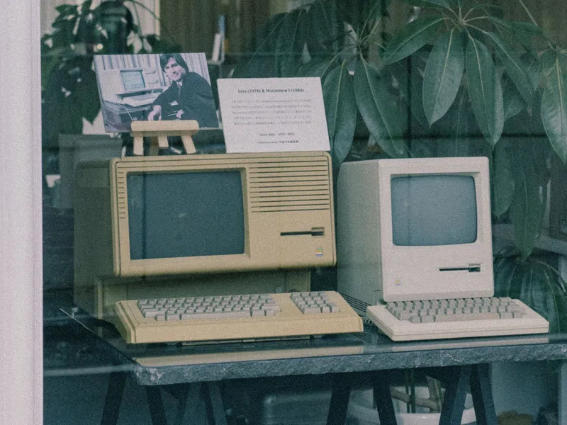 Two vintage personal computers displayed on a table, one beige and one cream, with a photo and plant background.