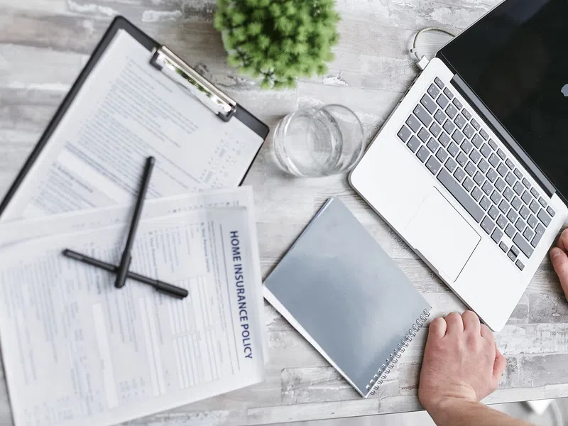Person working at a desk with a laptop, home insurance policy documents, notebook, and glass of water.