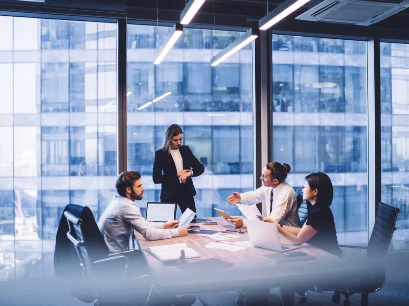 Business team discussing documents and laptops in modern office with cityscape view through glass windows.