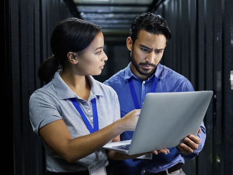 Two IT professionals discussing data on a laptop inside a server room with network racks in the background.