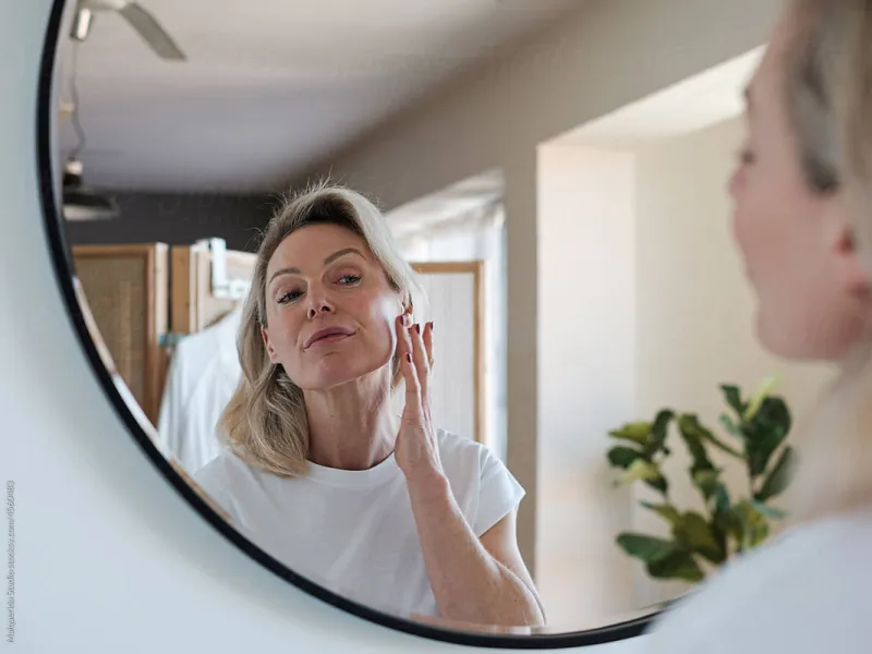 Mature woman applying skincare cream on her face while looking in a round mirror in a bright room