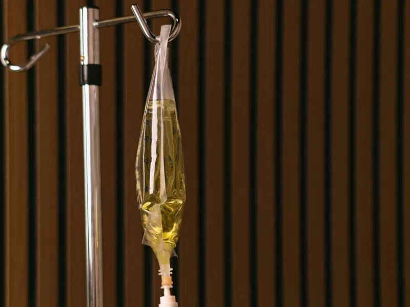 IV drip bag filled with yellow fluid hanging on a metal stand against a wooden slat wall background.