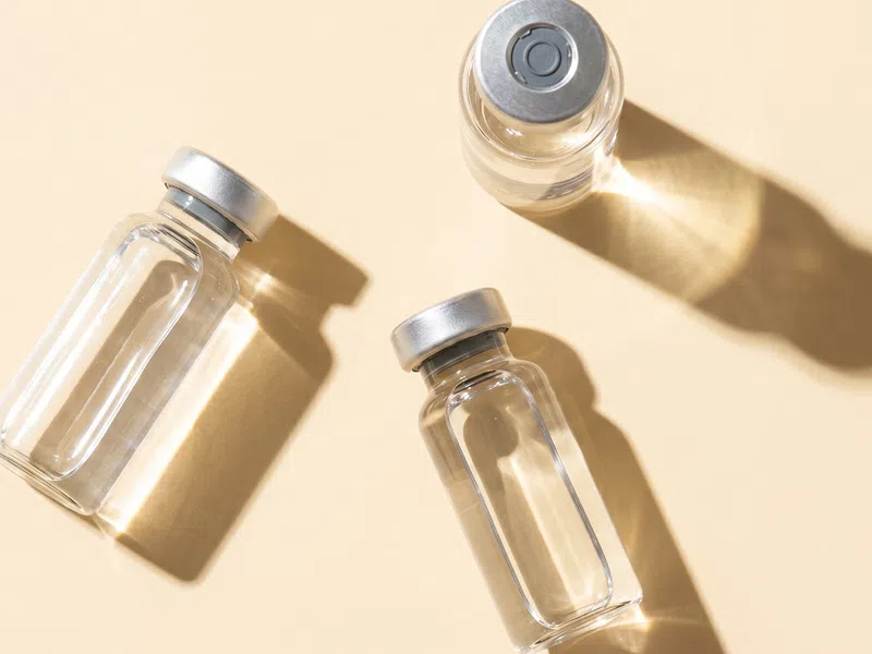 Three empty clear glass vials with silver caps casting shadows on a beige surface under bright light.
