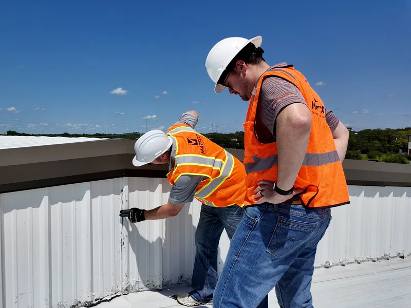 Two Mattco roofing technicians in white hard hats and orange safety vests inspecting the parapet wall and flashing detail on a commercial metal roof in Texas. One crouches to examine the wall base while the other supervises the inspection under a clear blue sky.