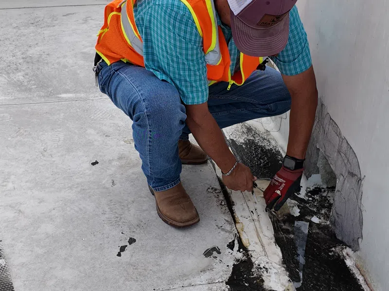 Mattco roofing technician in an orange safety vest and work gloves examining a damaged roof seam and deteriorated flashing detail along a parapet wall during a commercial roof repair assessment in Texas, exposing the underlying membrane and insulation.