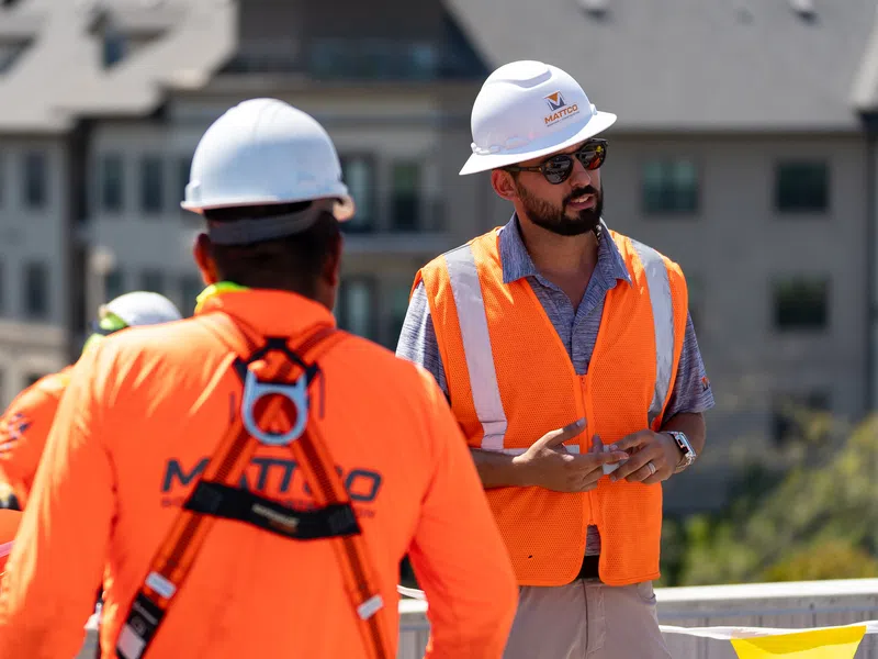 Mattco project manager in a white hard hat and orange safety vest directing crew members on a multi-family roofing project in Austin Texas with a large residential apartment complex visible in the background.