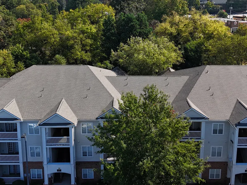 Aerial view of a modern three-story residential building surrounded by trees and parking areas under clear skies.