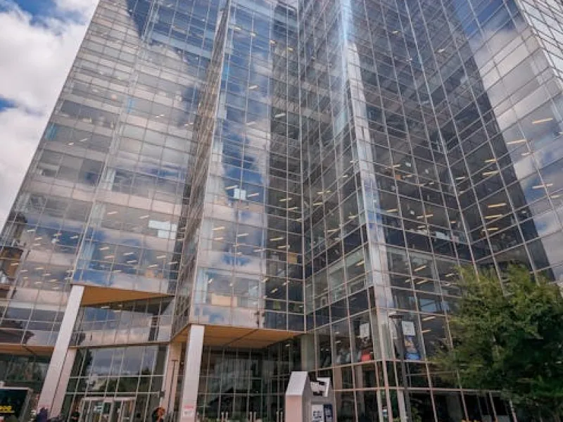 Modern glass office building with reflective windows, greenery, and partly cloudy sky overhead.