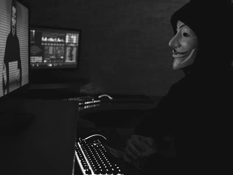 Person in Guy Fawkes mask and hoodie in dark room facing computer screen with hacker visuals.