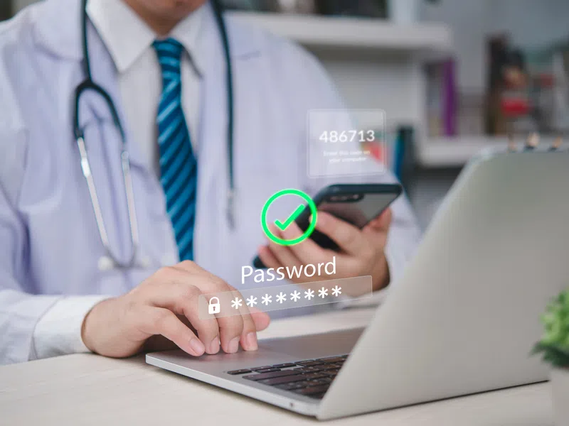 Doctor in lab coat using phone and laptop with secure password and two-factor authentication prompt.