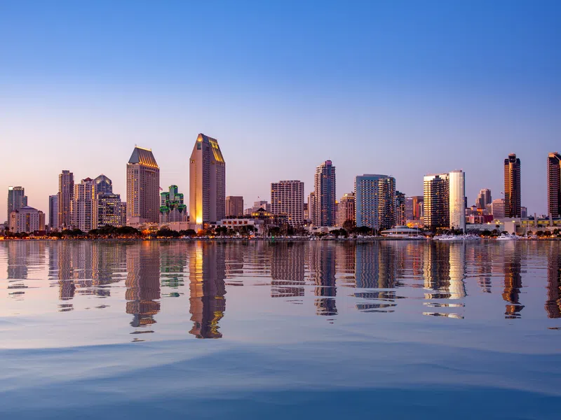 San Diego city skyline at sunset with buildings reflecting on calm harbor water and clear blue sky.