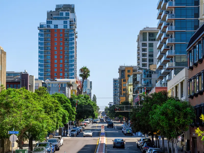 Urban street lined with green trees, parked cars, and modern high-rise buildings on a clear sunny day.