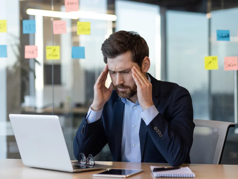 Stressed businessman in suit sitting at desk with laptop and sticky notes on glass wall in office.