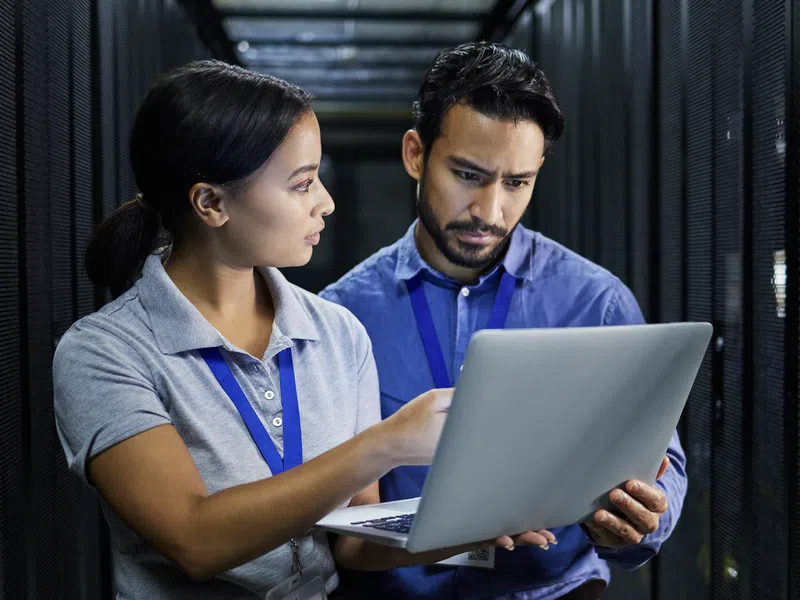Two IT professionals examining data on a laptop in a server room with racks of equipment around them.