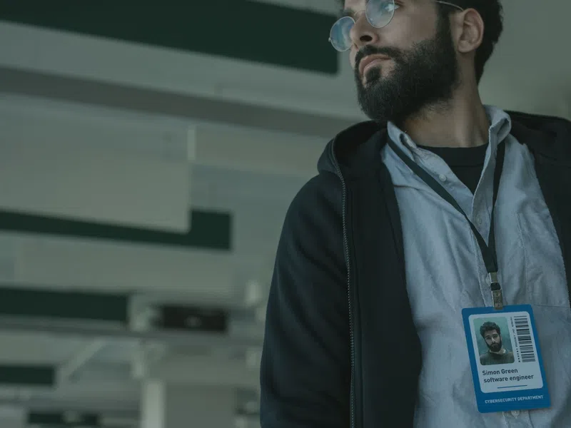 Bearded man wearing glasses and ID badge in an office setting looking to the side.