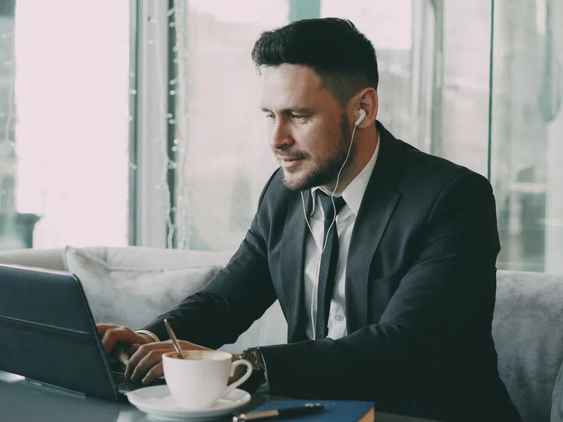 Man in business suit working on laptop with earphones at cafe table with coffee cup and notebook.