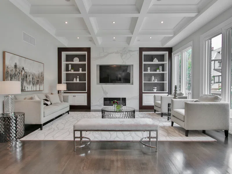 Modern living room with beige sofa, armchairs, marble accent wall, wall-mounted TV, large windows, and dark hardwood floors