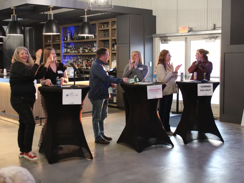 People socializing and clapping around standing cocktail tables at an indoor event with bar lighting.