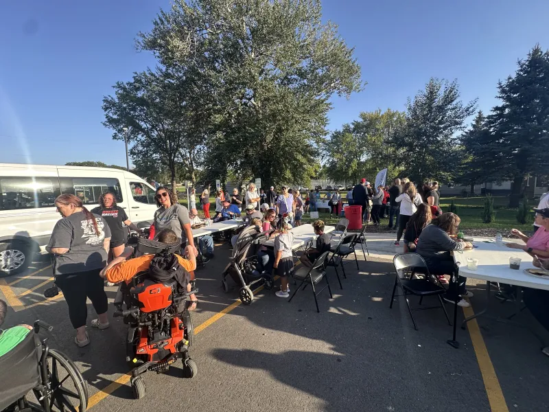 Community outdoor gathering with diverse people, tables, and wheelchairs under clear sunny sky and large trees.