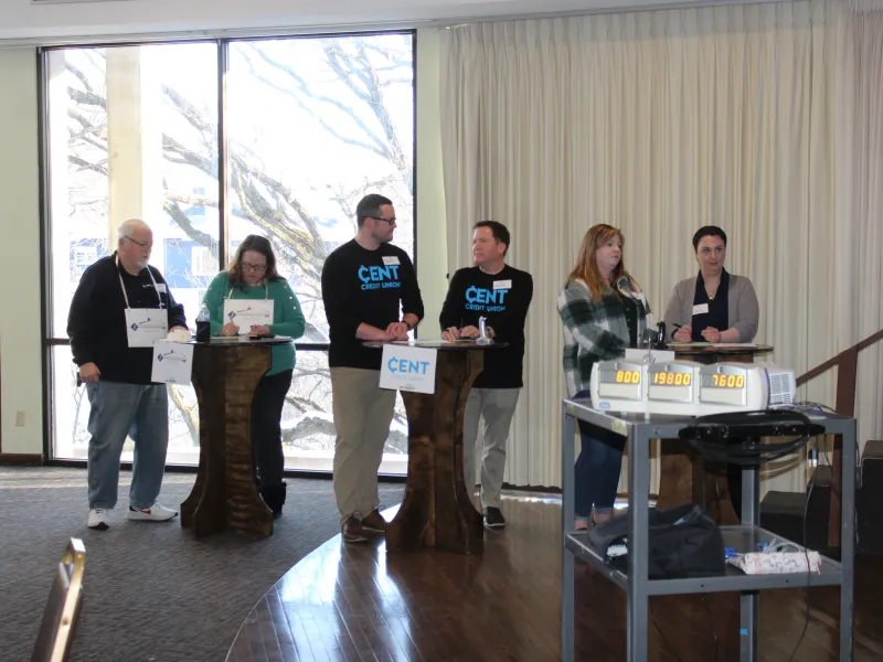 Six adults standing behind podiums in a room with large windows and a scoreboard on a cart.